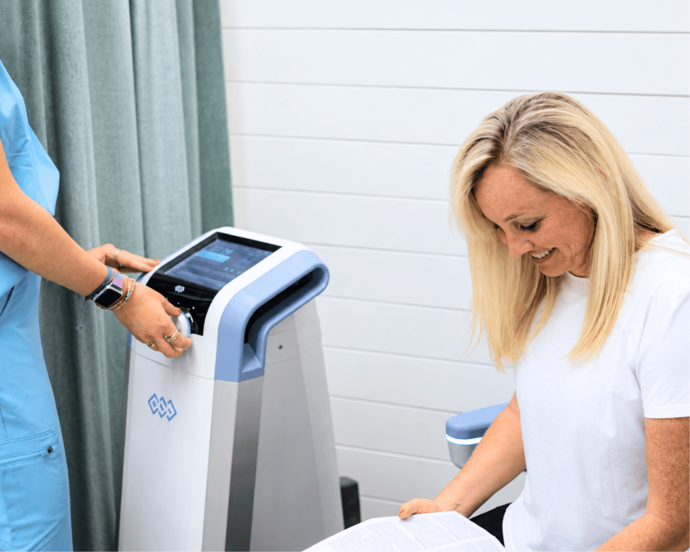 A woman smiles while a clinician operates a medical treatment device in a clinical setting. The woman holds paperwork and looks toward the device.