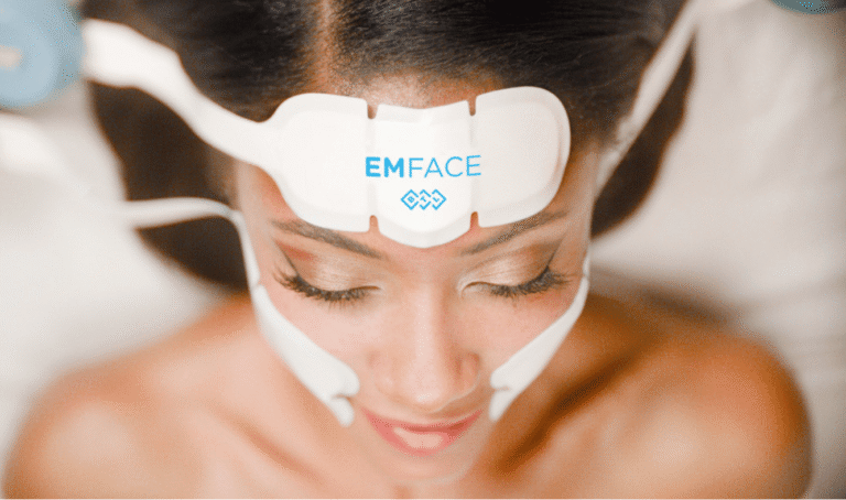 A woman with closed eyes receives an EMFACE facial treatment using white applicators positioned on her forehead and cheeks.