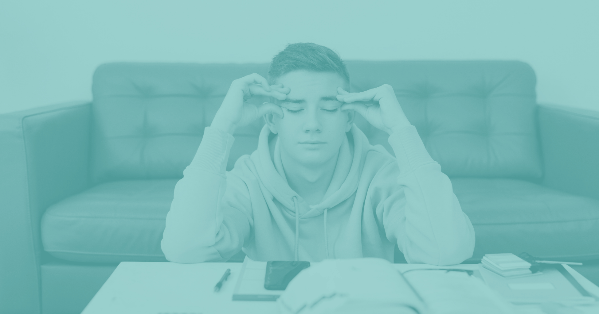 Young man sitting at a table with his eyes closed and hands at his temples, appearing stressed or overwhelmed.